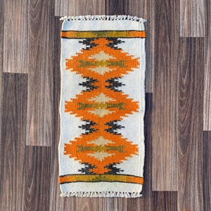 Op de afbeelding: Een rechthoekig geweven vloerkleed met een herhalend geometrisch patroon in oranje, zwart en beige op een witte achtergrond. Het kleed heeft een gefranjerde rand en ligt op een houten ondergrond.