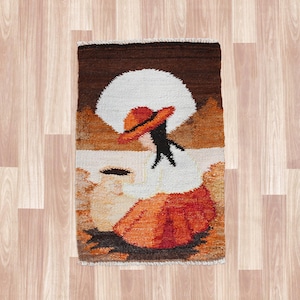 May include: A woven tapestry rug featuring a stylized figure in a hat and dress. The rug has a brown, orange, white, and black color scheme. The figure is depicted with a large white hat and an orange dress. The rug is rectangular.