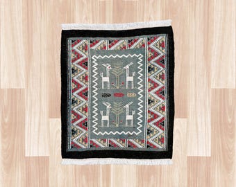 May include: A rectangular woven rug featuring a central sage green panel with white deer and tree motifs. The border displays a geometric pattern in red, black, and beige, framed by a solid black edge and finished with white fringe on both ends.