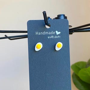 May include: A pair of handmade egg-shaped stud earrings. Each earring features a white egg white with a yellow yolk. The earrings are displayed on a dark gray card with the text "Handmade with love."