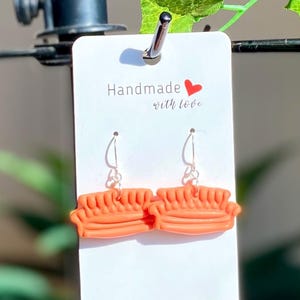 Op de afbeelding: Een paar oranje kleioorbellen in de vorm van een bank, hangend aan een wit kaartje met de tekst "Handmade with love" en een rood hart.