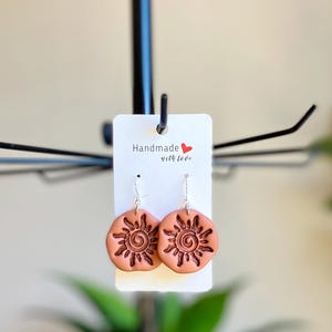 May include: A pair of terracotta sun earrings with a silver hook. The earrings are handmade and feature a spiral design in the center of the sun. The earrings are displayed on a white card with the text "Handmade with love".