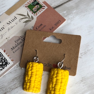 May include: A pair of handmade earrings shaped like yellow corn on the cob. The earrings have silver-colored hooks and are displayed on a brown card with the text "Handmade with love". A business card is in the background.