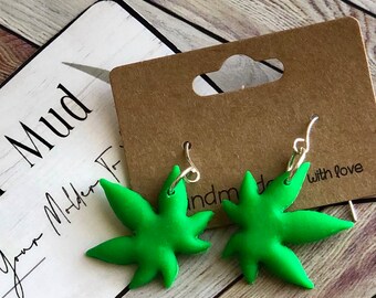 May include: Bright green leaf-shaped earrings with silver hooks. The earrings are displayed on a brown card with the words "handmade with love". The background is a light-colored wooden surface.