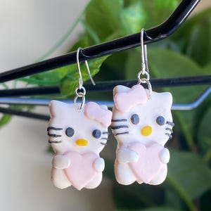Puede incluir: Un par de pendientes colgantes con un diseño de gato de dibujos animados blanco con bigotes negros, ojos negros y nariz amarilla. Cada pendiente tiene un corazón rosa y un lazo rosa. Los pendientes están suspendidos de ganchos plateados.