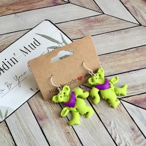 May include: A pair of green and purple bear-shaped earrings with silver hooks. The earrings are handmade with love.