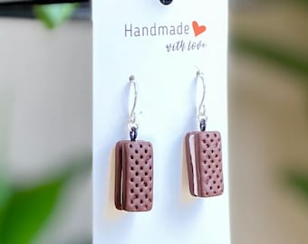 Puede incluir: Un par de pendientes de aro de plata con un dije de arcilla hecho a mano con forma de galleta de sándwich de chocolate. Los pendientes se muestran en una tarjeta blanca con el texto "Handmade with love".