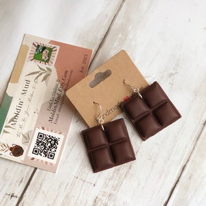 May include: A pair of chocolate bar earrings. The earrings are made of brown polymer clay and are shaped like a chocolate bar with four squares. The earrings are hanging from silver hooks.
