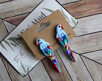 May include: Two handmade parrot-shaped earrings. Each earring features a light blue head, a colorful body with red, blue, green, and yellow accents, and a red tail. The earrings are displayed on a brown card with a hole for hanging.