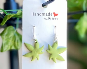 May include: Handmade earrings featuring a light green cannabis leaf design. The earrings are suspended from silver-colored hooks. The tag reads "Handmade with love."