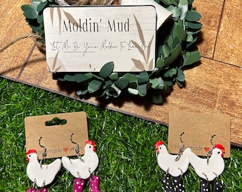 May include: Handmade chicken earrings with red combs and pink polka dot rain boots. The earrings are on a brown card with the word "handmade" and a heart. A sign that says "Moldin' Mud" is in the background.