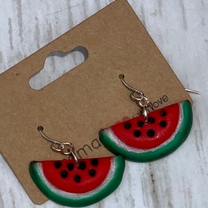 Può includere: Orecchini a forma di fetta di anguria con ganci color argento. Gli orecchini presentano un centro rosso con semi neri, una buccia verde e un bordo glitterato argentato. Sono esposti su una carta marrone.