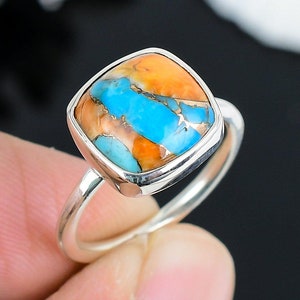 Puede incluir: Un anillo de plata con una piedra turquesa de forma cuadrada. La piedra tiene una mezcla de colores azules, naranjas y blancos.