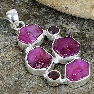May include: A silver pendant featuring irregularly shaped, vibrant pink gemstones. The pendant has a cluster design with smaller, round, dark red stones interspersed among the larger pink stones. The pendant is set against a textured gray surface.