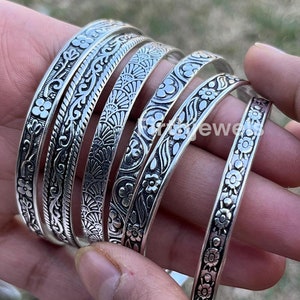 May include: Several silver bangles with detailed floral and patterned designs. The bangles are displayed on a person's wrist, highlighting the intricate craftsmanship and variety of styles. The image showcases the jewelry's texture and shine.