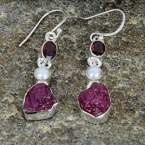 May include: A pair of silver dangle earrings with a red ruby and white pearl accents. The earrings feature a rough, natural ruby gemstone.