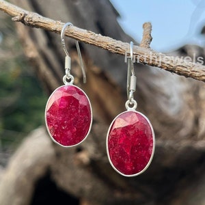 May include: A pair of silver earrings featuring oval-shaped red ruby gemstones. The earrings have a simple design and are ideal for everyday wear.