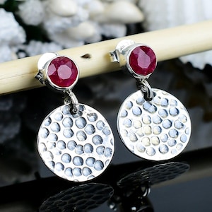 Może przedstawiać: Srebrne kolczyki sztyfty z teksturowanym wzorem tarczy. Każdy kolczyk ma mały, okrągły, ciemnoczerwony kamień szlachetny i większy, teksturowany dysk. Kolczyki są prezentowane na rozmytym tle.