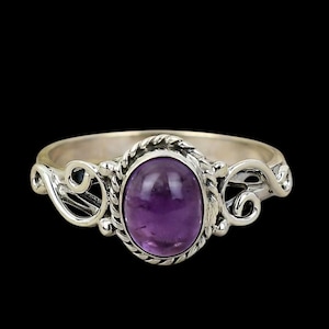 May include: A silver ring featuring a deep purple oval amethyst gemstone. The ring has a decorative, swirling design on the band and around the gemstone. The ring is set against a black background.