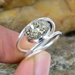 Op de afbeelding: Een zilveren ring met een grote, gefacetteerde pyriet steen in het midden. De ring is ontworpen met een dubbele band, wat een unieke en moderne uitstraling geeft.