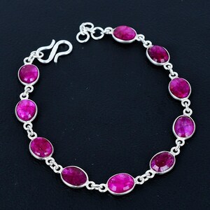 May include: A silver bracelet with oval, bright pink gemstones. The gemstones are connected by a delicate silver chain and fastened with a hook clasp. The bracelet is shown against a dark background.
