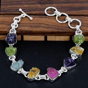 May include: A silver bracelet with a toggle clasp features seven different rough gemstones in a rainbow colour scheme. The gemstones are amethyst, peridot, aquamarine, citrine, ruby, peridot, and citrine.