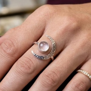Peut inclure: Une bague en argent avec un croissant de lune et une pierre précieuse ronde rose pâle. La lune a une texture martelée. Une petite bague en or avec de petites pierres claires est également visible.