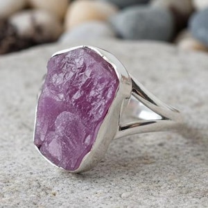 May include: A silver ring with a large, rough-cut, purplish-pink gemstone. The stone has a textured surface and is set in a simple, polished silver band. The ring is displayed on a light grey surface with a blurred background of rocks.