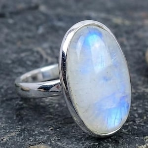 May include: A silver ring with a large, oval moonstone. The moonstone displays a milky white hue with iridescent blue flashes. The ring's band is polished silver.