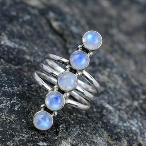 May include: A silver ring with five round, iridescent moonstone cabochons set in a vertical line. The ring has multiple bands that wrap around the finger. The moonstones have a blue and white colour.