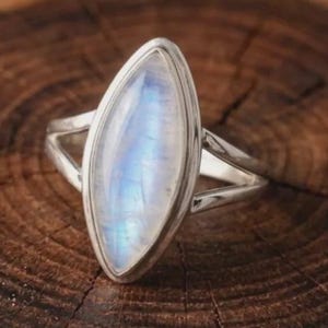 May include: A silver ring featuring a marquise-cut moonstone with a translucent, iridescent blue sheen. The gemstone is set in a smooth metal bezel, and the band consists of two thin, polished silver wires that meet at the base of the setting.