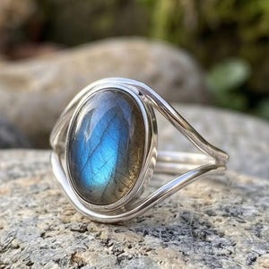 May include: A silver ring featuring an oval-shaped labradorite gemstone with a prominent blue iridescent flash. The stone is set in a smooth metal bezel, and the band consists of two thin, polished silver wires that split and rejoin at the sides.