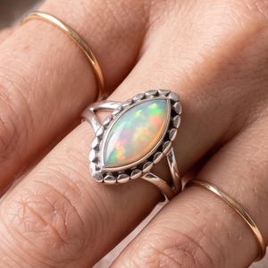 May include: A silver ring with an oval opal gemstone. The opal displays a range of colors, including white, yellow, and green. The ring has a detailed, beaded border and is worn on a finger.