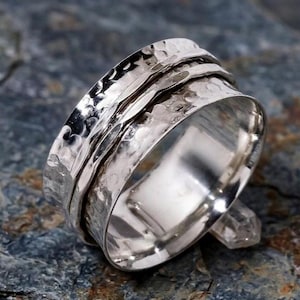 May include: A silver spinner ring with a textured, hammered finish. The ring has a wide band with a central spinning element. The ring is displayed on a dark, textured surface. The ring's design offers a tactile and stylish appeal.