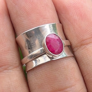 May include: A silver ring with a wide band and a red gemstone set in the centre. The ring is worn on a finger.