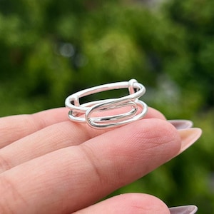 Puede incluir: Un anillo de plata con un diseño abstracto único. El anillo está hecho de tres bandas entrelazadas, creando un estilo moderno y minimalista.