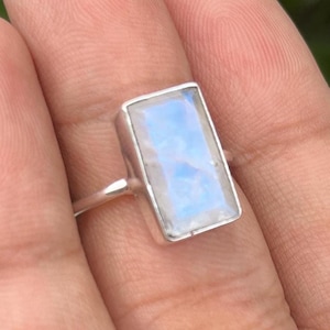 May include: A silver ring featuring a rectangular moonstone gemstone. The stone is milky white with a subtle iridescent shimmer.