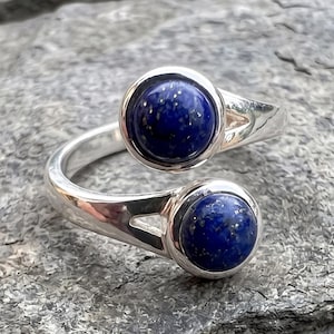 Op de afbeelding: Een open ring van gepolijst zilver, voorzien van twee ronde, diepblauwe lapis lazuli cabochons aan beide uiteinden. De stenen vertonen subtiele goudkleurige spikkels op hun gladde, afgeronde oppervlakken, wat contrasteert met de heldere, reflecterende metalen band.