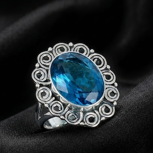 Könnte beinhalten: Ein silberner Ring mit einem großen, oval geschliffenen, leuchtend blauen Edelstein. Der Edelstein ist in eine dekorative Silberfassung mit einem wirbelnden, scrollartigen Design gefasst. Der Ring ist vor schwarzem Hintergrund platziert.