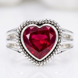 Puede incluir: Anillo de plata con una piedra preciosa roja oscura en forma de corazón. La piedra preciosa está engastada en un borde retorcido. El anillo tiene un diseño de triple banda. Joya romántica. Se muestra sobre una superficie blanca.