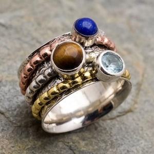 May include: A multi-band spinner ring featuring a wide silver base, a textured copper band, a hammered silver band, and a gold-toned beaded band. Three bezel-set gemstones are mounted on the rings: a dark blue lapis lazuli, a brown tiger's eye, and a light blue topaz.