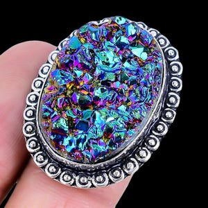 May include: An oval-shaped, iridescent druzy gemstone ring. The gemstone displays a spectrum of colors, including blues, greens, purples, and yellows. The ring has a silver-colored, ornate setting with a detailed border. The ring is held by a person's finger.