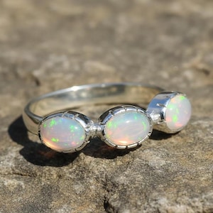 May include: A silver ring with three oval opal gemstones. The opals show a play of colour with green and red flashes. The ring is set on a natural stone surface.