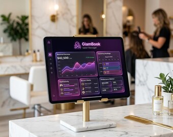 May include: A tablet displaying the "GlamBook Salon Manager" app, mounted on a gold stand with a marble base. The screen shows revenue and performance metrics. A gold comb and perfume bottles are on the marble countertop.