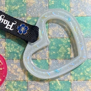 May include: Two heart-shaped resin keychains with a black ribbon with the text "Harajuku" and "Kyoto" on it. The hearts are filled with glitter and have a floral design.