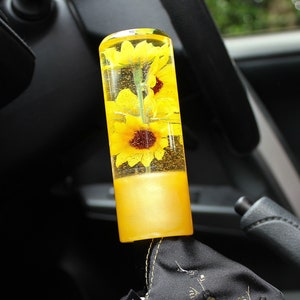 May include: A custom car gear stick knob featuring sunflowers set in clear resin. The knob has a yellow base and is paired with a black shift boot with a gold floral pattern. The shift boot has the text "HAYAKUCO".