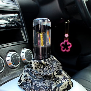 May include: A black leather car interior with a custom gear shift knob. The knob is made of clear acrylic and has a gold-coloured design inside. The knob is covered with a black and gold fabric boot with a dragon pattern.