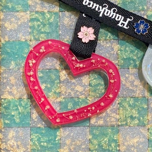 May include: Two heart-shaped resin charms with floral details. One charm is pink with white flowers and the other is white with blue flowers. The charms are attached to a black ribbon with the text "Harajuku" and "kuco" printed on it.