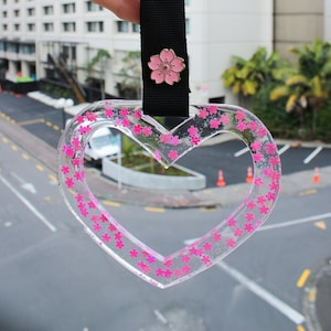 Puede incluir: Un llavero transparente en forma de corazón con purpurina rosa y un pequeño dije de flor rosa. El llavero está unido a una correa negra con la palabra "Hauyakuko" impresa en blanco.