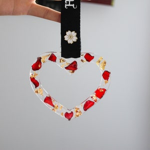 May include: A clear heart-shaped ornament with red and gold accents. The ornament is hanging from a black strap with the text "Hayakkuco" printed in white. A white flower with a gold centre is attached to the strap.
